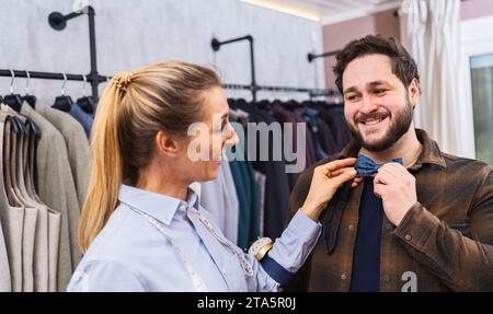 Saleswoman fitting a bow tie on a customer, both are smiling, racks of clothes behind in a men's clothing store Stock Photo