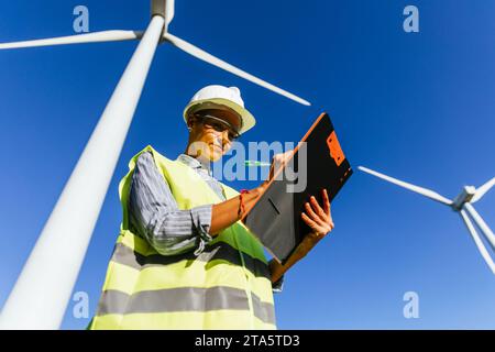 Woman engineer writing on a clipboard against a wind farm. Stock Photo