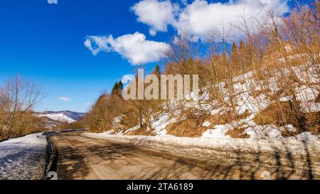 old road pass through snow covered hills. mountainous winter landscape on a sunny day beneath a blue sky with clouds Stock Photo