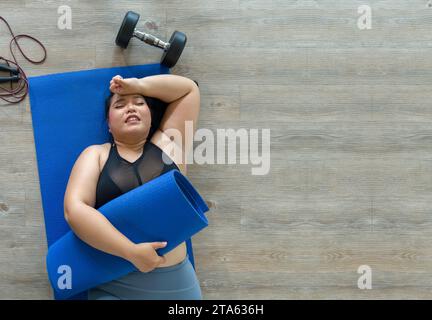 A plus-size woman lies exhausted on a blue mat, taking a breather after a vigorous workout session with dumbbell and jump rope. Top view Stock Photo