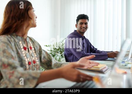 Smiling Indian businessman talking to coworker sitting next to him on office Stock Photo