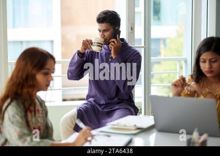 Indian businessman in purple kurta drinking coffee and talking on phone with colleague Stock Photo