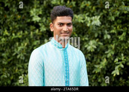 Smiling Indian young man in traditional kurta standing against green fence Stock Photo