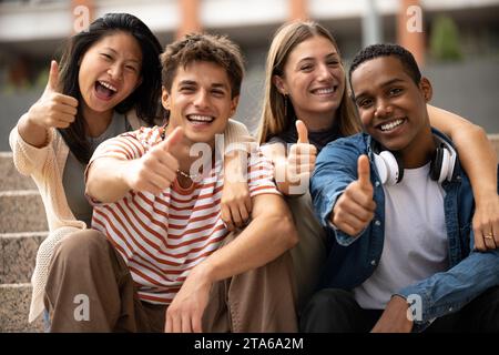 Cheerful multicultural group of friends showing thumbs up sitting on stairs. Happy young students smiling and satisfied sitting outside. Stock Photo