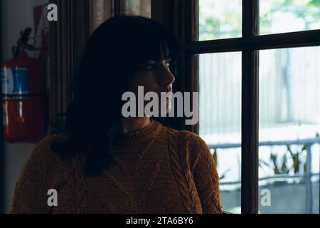 young woman in dark is thoughtful and worried indoors looking outside through door window, copy space Stock Photo