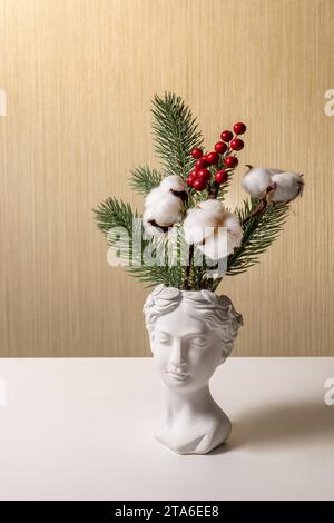Vase in form of an aphrodite's head with bouquet of spruce, cotton and berries stands on table. The concept of facial care in winter. Stock Photo