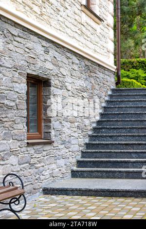 Granite steps along the stone wall of the house with a window lead to the green garden in the background. Vertical image. Stock Photo