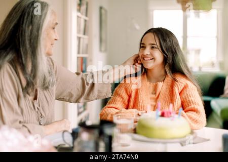 Senior woman touching cheek of granddaughter sitting with birthday cake at dining table Stock Photo