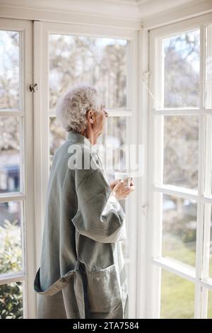 Contemplative senior man holding coffee cup while looking through window at home Stock Photo