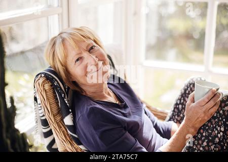 Portrait of smiling blond senior woman with coffee cup sitting on chair at home Stock Photo