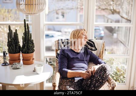 Senior woman with eyes closed listening music through in-ear headphones while relaxing on chair at home Stock Photo
