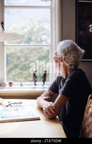 Senior man looking through window while sitting at home Stock Photo