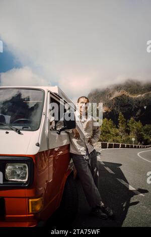Portrait of smiling young man standing next to van on road during road trip Stock Photo
