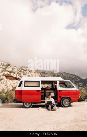 Young man using laptop while sitting near van against sky Stock Photo