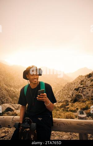 Smiling young man using mobile phone while sitting on railing against sky Stock Photo