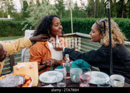 Man and woman looking at necklace of female friend sitting at dining table in back yard Stock Photo