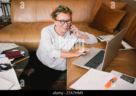 Happy senior woman talking on mobile phone and using laptop at home Stock Photo