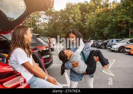 Girl sitting in trunk looking at mother having fun with daughter near car Stock Photo