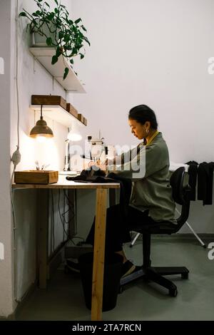Side view of seamstress stitching clothes using sewing machine while sitting on chair at workshop Stock Photo