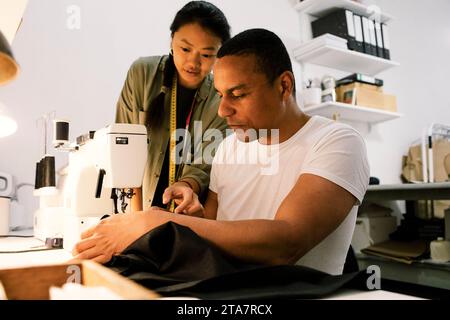 Female colleague assisting male tailor stitching clothes using sewing machine at workshop Stock Photo