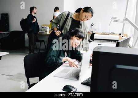 Female technician repairing laptop while sitting near customer at store Stock Photo