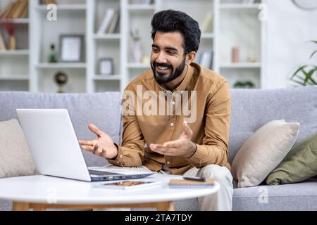 Happy handsome young indian man freelancer talking by online video call conference meeting on laptop computer having remote work or distance education learning on couch at home High quality photo Stock Photo