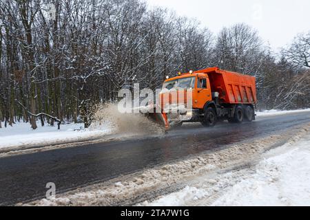 A large car with a plow clears the road from snow. Orange cargo special equipment is struggling with the elements in winter. Removing the effects of t Stock Photo