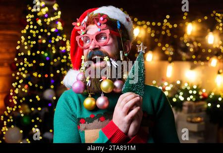 Happy Santa man with New Year balls in beard holds small Christmas tree. Christmas beard style. Bearded guy in party glasses and Santa hat with little Stock Photo