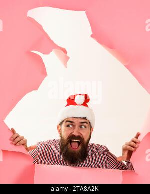 Happy bearded man in Santa hat looking through paper hole. Smiling guy in checkered shirt making hole in paper. Winter holidays and boxing day Stock Photo