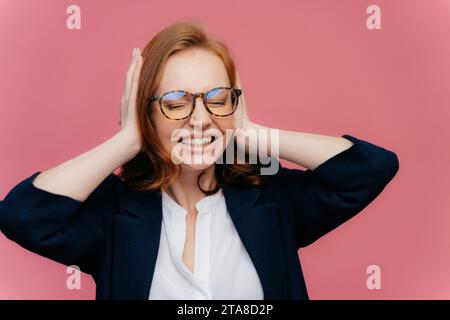 Joyful businesswoman with hands on head, laughing, in stylish eyewear, blue blazer over white shirt, pink backdrop Stock Photo