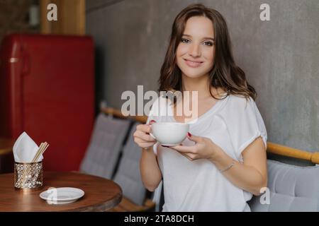 Content woman enjoying a cup of coffee in a cafe, relaxed atmosphere, white t-shirt, and a warm, inviting smile Stock Photo