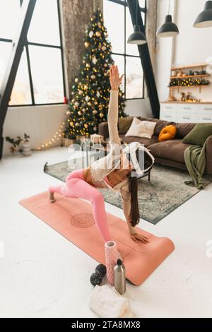 In her sports gear and a virtual reality headset, the trendy woman practices yoga near a Christmas tree. Stock Photo