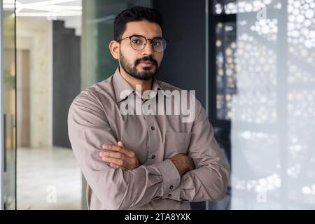 Portrait of serious confident hindu businessman, man in shirt thinking and looking at camera with crossed arms, mature boss working inside office at workplace, financier accountant in glasses. Stock Photo