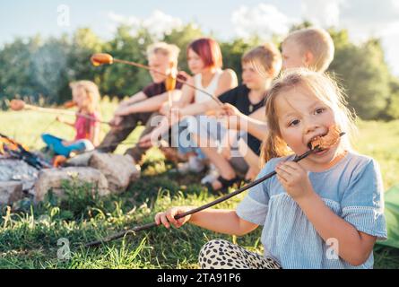 Portrait of little girl eating grilled sausage. Group of Kids - Boys and girls roasting sausages on long sticks over a campfire flame. Outdoor active Stock Photo