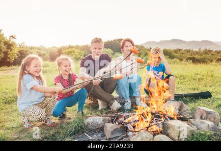 Five kids group Boys and girls cheerfully laughed and roasted sausages on sticks over a campfire flame near the green tent. Outdoor active time spendi Stock Photo