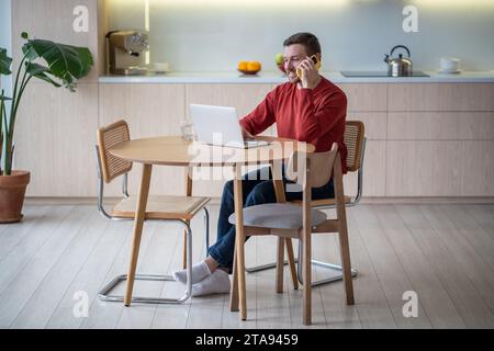 Smiling man working from home as freelancer using laptop, speaking on smartphone during work break Stock Photo