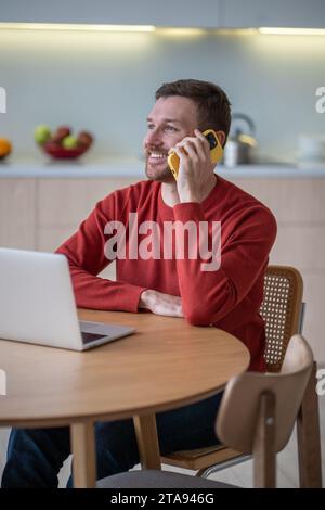 Digital nomad. Pleased man freelancer smiling talk cellphone call remote working with laptop at home Stock Photo