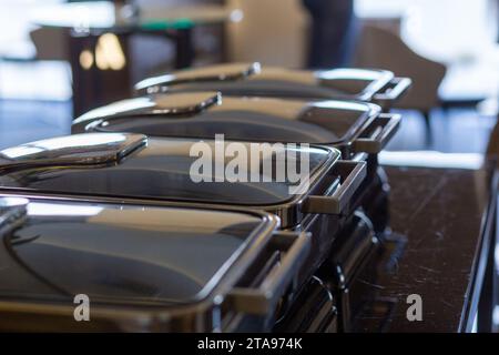 Row of closed buffet food dishes at party banquet hall. Chafing Dish ready for service made of stainless steel at buffet. Stock Photo