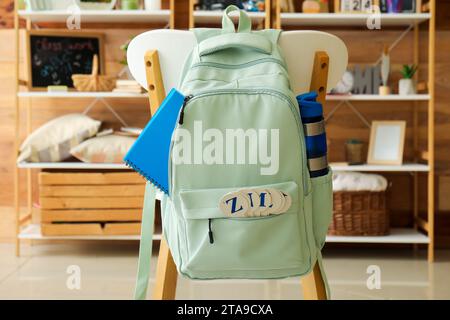 Stylish school backpack with bottle of water and stationery hanging on chair in room Stock Photo
