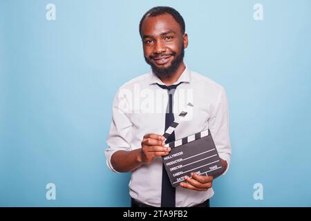 African American movie director holding open clapperboard in hands looking confident while posing for camera. Young successful filmmaker grasping clapper standing proud against blue background. Stock Photo