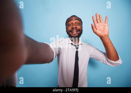 On blue background, cheerful vlogger waves hello to the camera during videocall conference. Point of view of african american influencer taking a selfie while making hand gesture for social media. Stock Photo