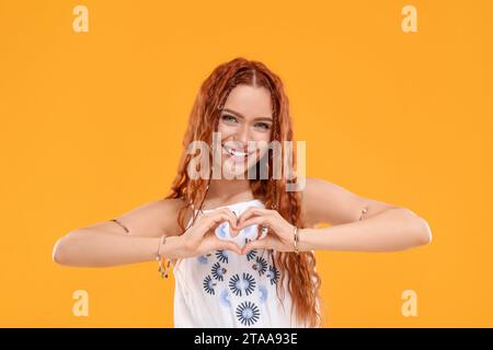 Beautiful young hippie woman making heart with hands on orange background Stock Photo