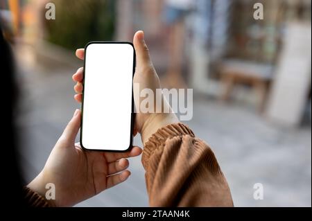 A woman holding a white-screen smartphone mockup over a blurred background of a city. A woman using her smartphone while walking in the city. Stock Photo