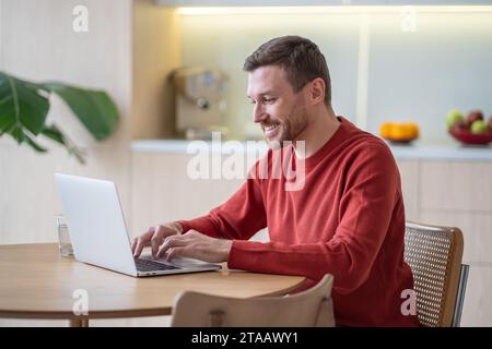 Positive cheerful man enjoying work from home as freelancer using laptop in cozy modern apartment Stock Photo