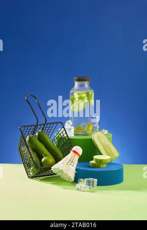 Cucumber detox water bottle standing on a podium, displayed with a shuttlecock and a basket of fresh cucumbers. Concept of healthy organic fruit Stock Photo