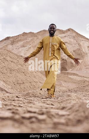 Vertical full length portrait of young Black man wearing traditional clothing and walking towards camera in desert landscape Stock Photo