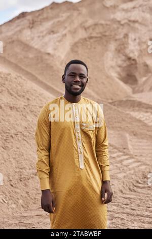 Vertical portrait of Black young man wearing traditional kaftan dress walking towards camera in desert Stock Photo