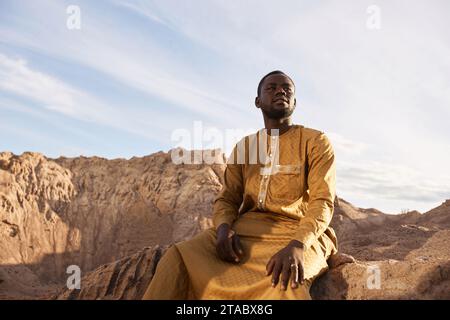 Dramatic portrait of Black young man wearing long kaftan sitting sand dune and looking at sky in desert, copy space Stock Photo