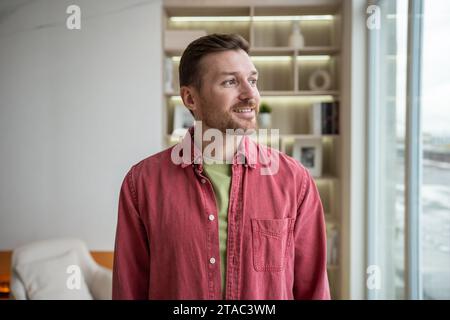 Smiling relaxed man looking at window in new home apartment thinking about future, daydreaming. Stock Photo