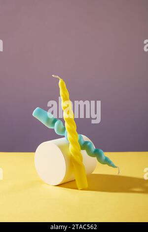 Two twisted candles in two colors: yellow and blue are placed next to a white podium on a yellow table with a purple background. Creative space for de Stock Photo